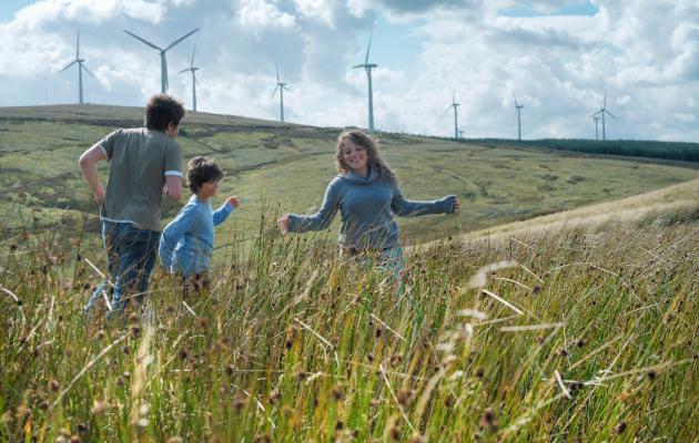 Young people in long grass in the foreground with wind turbines in the background.