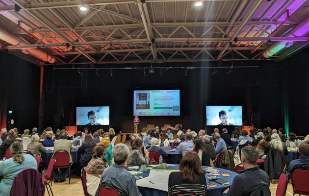 Image of tables with people sat around them in a large room with a big screen at the back 