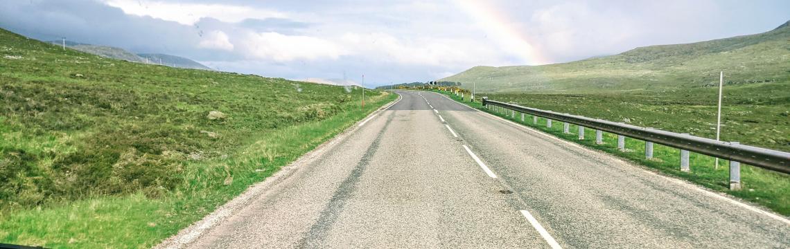 Open road in Scottish Highlands with a rainbow in the background.