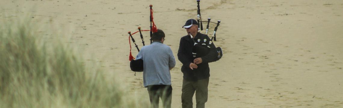 Two men standing on a beach playing bagpipes